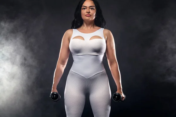 Taille plus femme sportive femme en tenue de sport blanche, athlète avec haltères rend l'exercice de remise en forme sur fond noir avec des lumières. Motivation pour les personnes grasses . — Photo