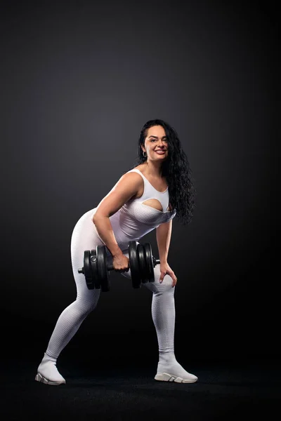 Talla más mujer en forma deportiva mujer en ropa deportiva blanca, atleta con mancuernas hace ejercicio de fitness sobre fondo negro con luces. Motivación para las personas gordas . — Foto de Stock