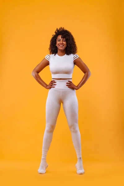 Улыбается сильная атлетичная женщина с черной кожей и вьющимися волосами, делает упражнения на жёлтом фоне в спортивной одежде. Фитнес и спортивная мотивация . — стоковое фото