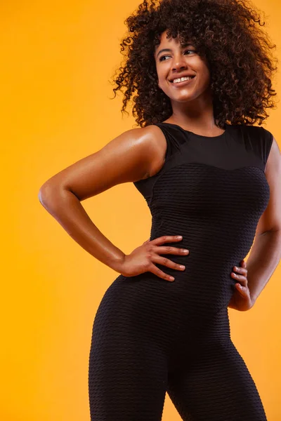 Sorridente donna atletica forte con pelle nera e capelli ricci, facendo esercizio su sfondo giallo indossando abbigliamento sportivo. Fitness e motivazione sportiva. — Foto Stock