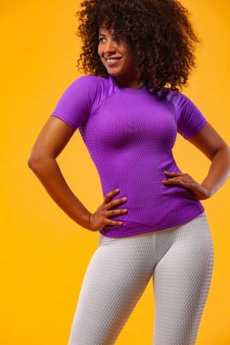 Gülümseyen, siyah tenli, kıvırcık saçlı, spor giyim kuşamında sarı arka plan üzerinde egzersiz yapan güçlü atletik kadın. Spor ve spor motivasyonu.