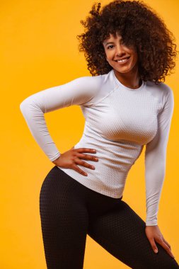 Gülümseyen, siyah tenli, kıvırcık saçlı, spor giyim kuşamında sarı arka plan üzerinde egzersiz yapan güçlü atletik kadın. Spor ve spor motivasyonu.