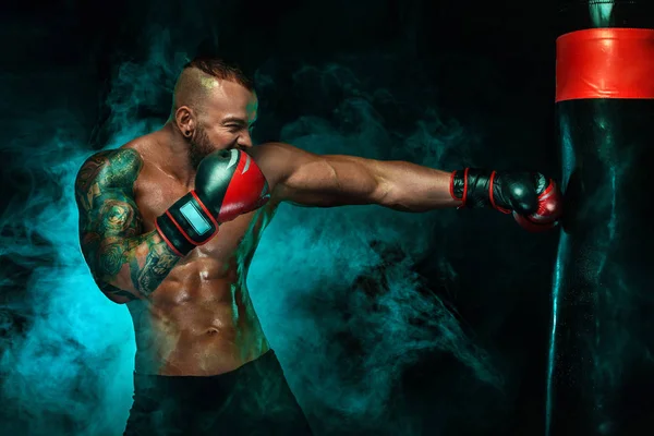 Sportoló a tetoválás, férfi bokszoló harcok a kesztyűt boksz boxzsák. Izolált fekete háttér füst. Lemezterület másolása. — Stock Fotó