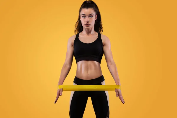 Mujer atleta deportiva musculosa joven en ropa deportiva negra con bandas o expansor en el gimnasio sobre fondo amarillo. Espacio de copia para anuncios de nutrición fitness . — Foto de Stock