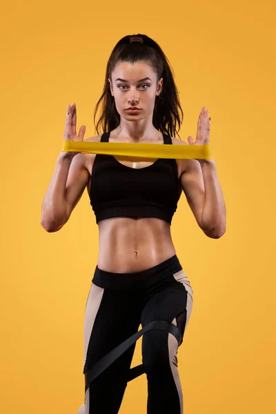 Mujer atleta deportiva musculosa joven en ropa deportiva negra con bandas o expansor en el gimnasio sobre fondo amarillo. Espacio de copia para anuncios de nutrición fitness . — Foto de Stock