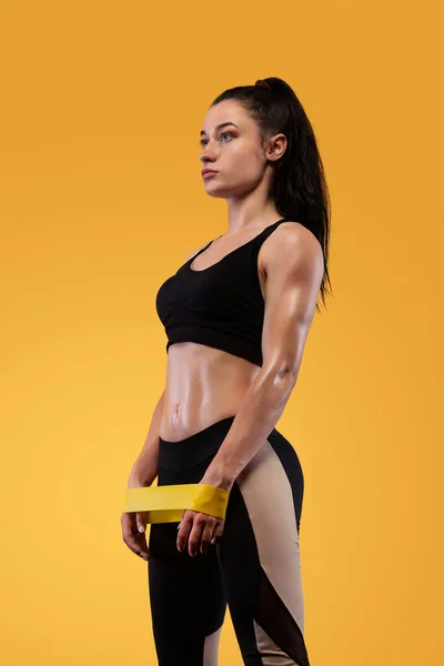 Mujer atleta deportiva musculosa joven en ropa deportiva negra con bandas o expansor en el gimnasio sobre fondo amarillo. Espacio de copia para anuncios de nutrición fitness . — Foto de Stock