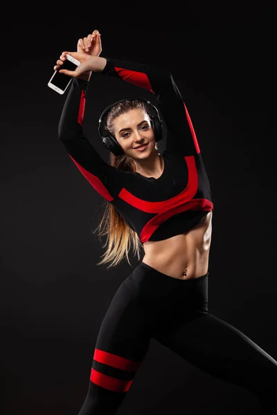 Hermosa mujer joven y en forma en ropa deportiva y auriculares escuchando música desde el teléfono y sonriendo. Aislado sobre un fondo negro . — Foto de Stock