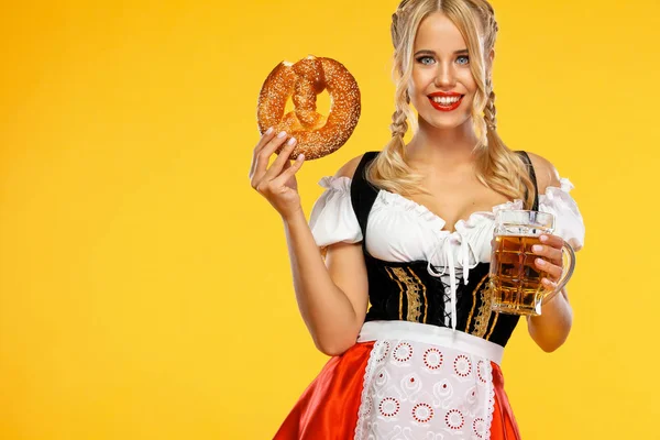 Jeune serveuse sexy Oktoberfest girl, vêtue d'une dirndl bavaroise ou allemande traditionnelle, servant de grandes tasses à bière avec boisson et bretzel, isolée sur fond jaune . — Photo