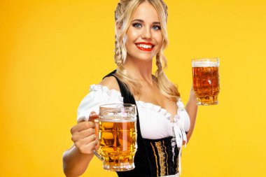 Geleneksel bir Bavyera ya da Alman dirndl 'i giymiş genç seksi Ekim Festivali kız garsonu, sarı arka planda içkili iki büyük bira bardağı servis ediyor..