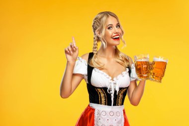 Geleneksel bir Bavyera ya da Alman dirndl giyip sarı arka planda izole edilmiş büyük bira bardakları servis eden genç seksi Ekim Festivali kız garsonu. Yukarıya bakan bir kadın.