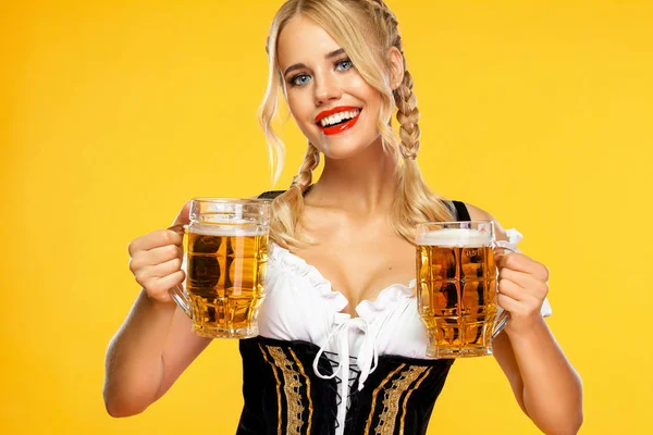 Młoda seksowna kelnerka Oktoberfest, ubrana w tradycyjny bawarski lub niemiecki dirndl, serwująca dwa duże kubki piwa z napojem odizolowanym na żółtym tle. — Zdjęcie stockowe