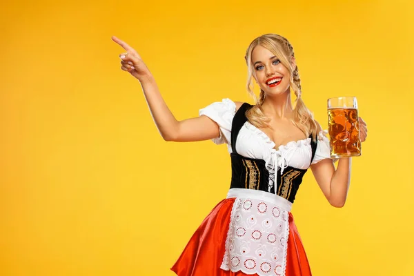 Mladá sexy servírka Oktoberfestu, oblečená v tradičním bavorském nebo německém dirndl, servíruje velké pivní džbánky s pitím izolovaným na žlutém pozadí. Žena ukazující doleva. — Stock fotografie