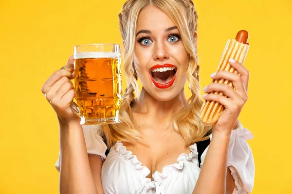 Młoda seksowna kelnerka Oktoberfest, ubrana w tradycyjny bawarski lub niemiecki Dirndl, serwująca duże kufle piwa z napojem i francuskim gorącym psem wyizolowanym na żółtym tle. — Zdjęcie stockowe