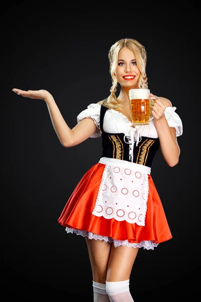 Lächelnde sexy Oktoberfestkellnerin, die ein traditionelles bayerisches oder deutsches Dirndl trägt und zwei große Bierkrüge mit Getränken auf schwarzem Hintergrund serviert. Frau zeigt nach links. — Stockfoto