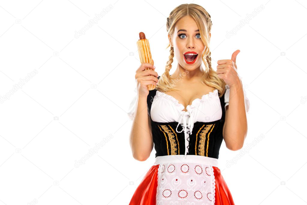 Young sexy Oktoberfest girl waitress, wearing a traditional Bavarian or german dirndl with french hot dogs isolated on white background. Wow emotion, woman with thumbs up.