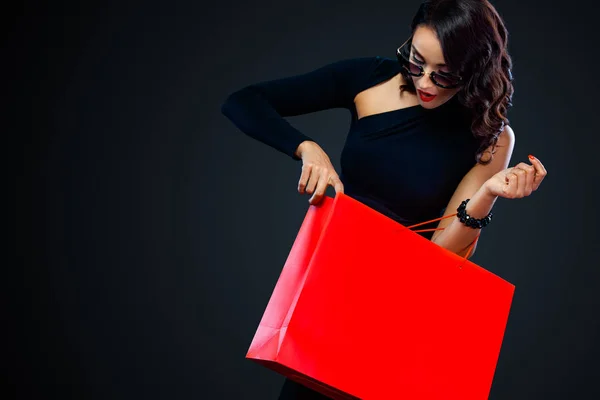 Venerdì nero concetto di vendita per negozio. Shopping donna in occhiali da sole in possesso di borsa rossa isolato su sfondo scuro . — Foto Stock