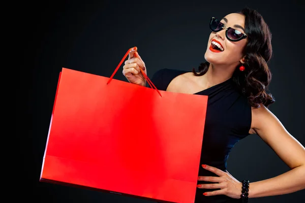 Conceito de venda Black Friday para loja. Menina sorridente feliz em óculos de sol segurando grande saco vermelho isolado no fundo escuro nas compras. Mulher apontando para olhar no espaço de cópia para texto de vendas . — Fotografia de Stock