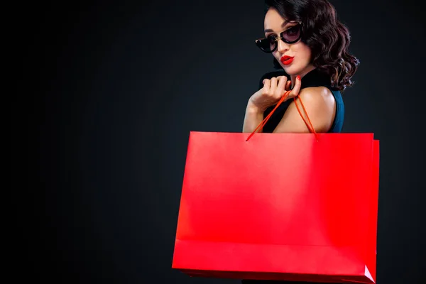 Black Friday sale concept for shop. Shopping woman in sunglasses holding red bag isolated on dark background. — Stock Photo, Image