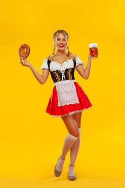 Genç seksi Oktoberfest kız garson, geleneksel Bavyera veya Alman dirndl giyen, içki ve bretzel ile büyük bira kupaları hizmet, sarı arka plan üzerinde izole. Tam boy fotoğraf