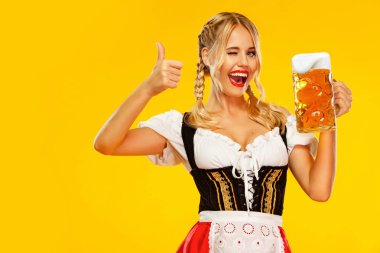 Vay duygu, başparmak yukarı kadın. Genç seksi Oktoberfest kız garson, geleneksel Bavyera veya Alman dirndl giyen, sarı arka plan izole içki ile büyük bira kupa hizmet.