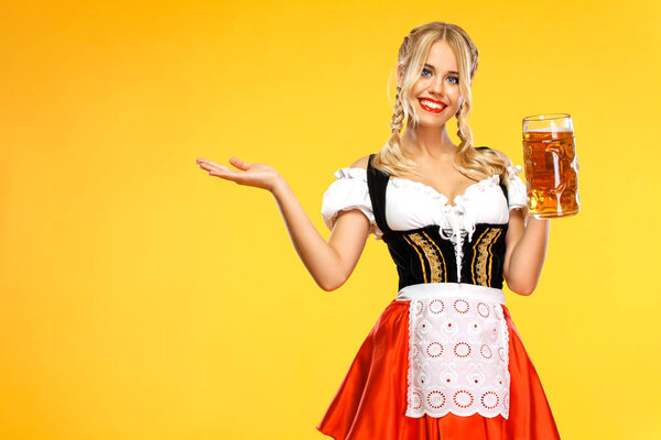 Young sexy Oktoberfest girl waitress, wearing a traditional Bavarian or german dirndl, holding big beer mug with drink isolated on yellow background. Woman pointing to looking left.