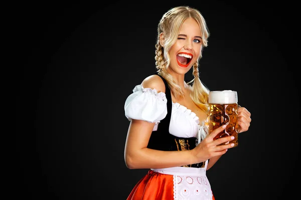 Souriant jeune fille sexy oktoberfest serveuse, portant un dirndl bavarois ou allemand traditionnel, servant une grande tasse de bière avec boisson isolée sur fond noir . — Photo
