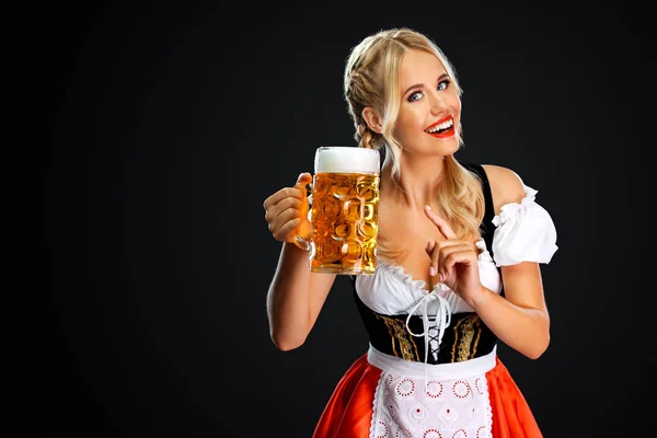 Lächelnde junge sexy Oktoberfestkellnerin, die ein traditionelles bayerisches oder deutsches Dirndl trägt und einen großen Bierkrug mit Getränken auf schwarzem Hintergrund serviert. — Stockfoto