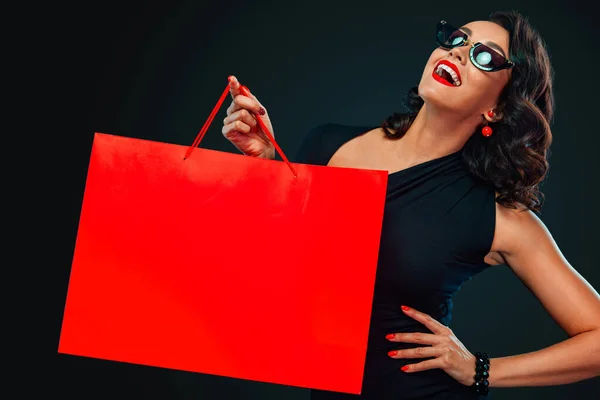 검은 금요일 쇼핑 컨셉이야. 쇼핑 할 때어두운 배경에 커다란 빨간 가방을 들고 선글라스를 쓴 행복하게 웃는 여자. 원문을 위한 복사 공간. — 스톡 사진