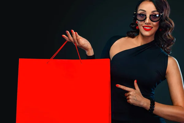 블랙 프라이데이 세일 컨셉의 숍. 어두운 배경에 고립 된 빨간 가방을 들고 선글라스에 쇼핑 소녀. 복사 공간에 왼쪽을 보고 가리키는 여자. — 스톡 사진