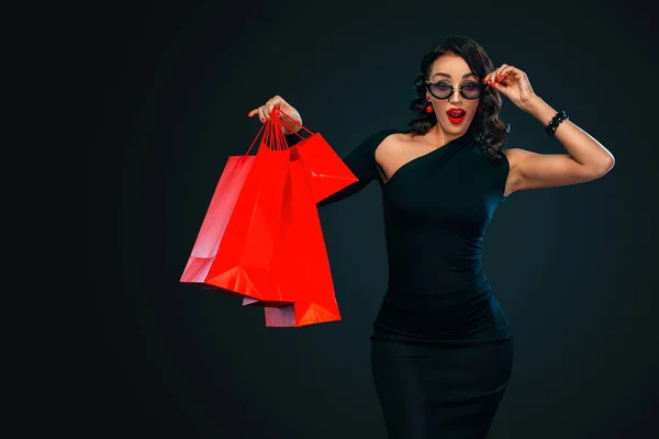 Conceito de venda Black Friday para loja. Mulher de compras em óculos de sol segurando saco vermelho isolado no fundo escuro . — Fotografia de Stock