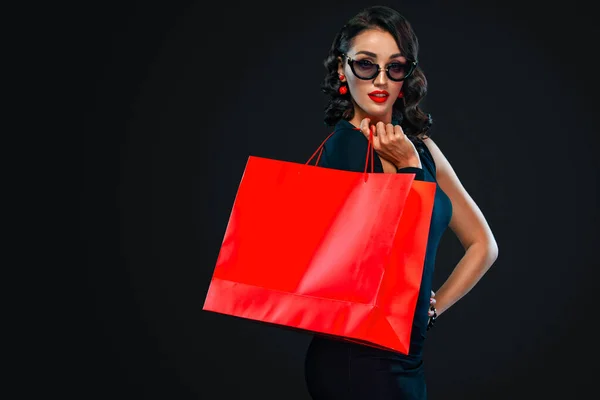 Conceito de venda Black Friday para lojas. Mulher de compras em óculos de sol segurando saco vermelho isolado no fundo escuro . — Fotografia de Stock