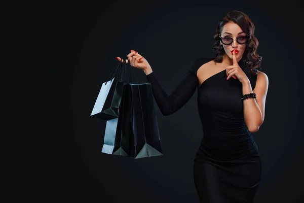 Black Friday concepto de venta para tienda. Chica en gafas de sol sosteniendo gran bolsa aislada sobre fondo oscuro en las compras. Mujer apuntando a buscar espacio de copia para texto de ventas . —  Fotos de Stock