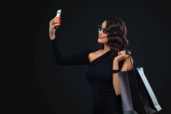 Black Friday és Cyber Monday eladó koncepció a bolt. Bevásárló nő mosollyal az arcán tartó táskát, és szelfit az okostelefonon. Sötét háttérrel elszigetelve — Stock Fotó