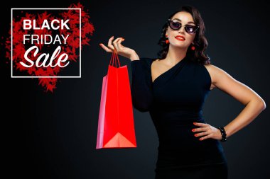 Dükkan için Black Friday satış konsepti. Alışverişte koyu arka planda izole büyük kırmızı çanta tutan güneş gözlüğü mutlu gülümseyen kız. Satış metni için kopya alanına bakmaya işaret eden kadın.
