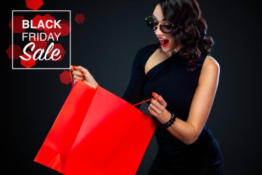 Dükkan için Black Friday satış konsepti. Alışverişte koyu arka planda izole büyük kırmızı çanta tutan güneş gözlüğü mutlu gülümseyen kız. Satış metni için kopya alanına bakmaya işaret eden kadın.