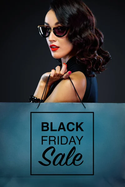 Closeup portrait of black friday sale concept for shop. Woman in sunglasses holding big bag isolated on dark background at shopping. — Stock Photo, Image