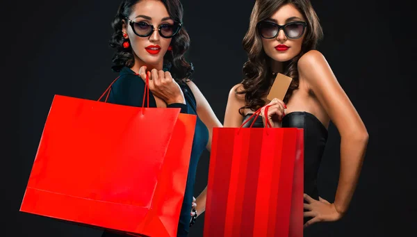 Black Friday concept de vente pour les magasins. Deux femmes en lunettes de soleil tenant sac rouge isolé sur fond sombre au centre commercial . — Photo