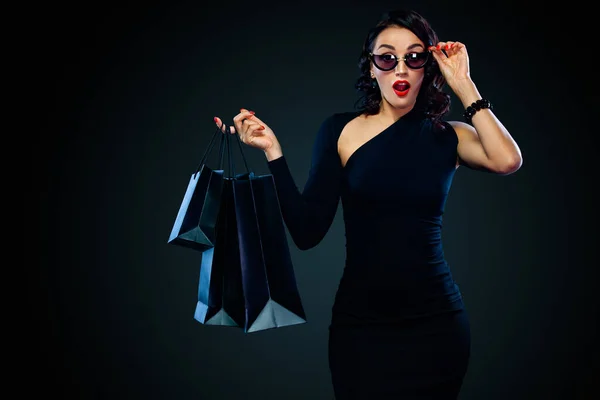 Black Friday sale concept for shop. Girl in sunglasses holding big bag isolated on dark background at shopping. Woman pointing to looking on copy space for sales text. — Stock Photo, Image