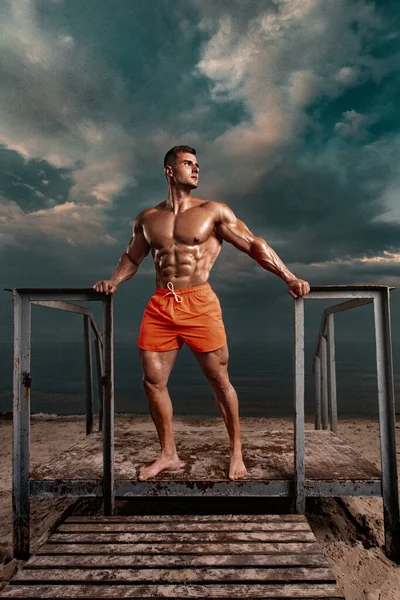 Coupe bodybuilder athlète sur la plage. Jeune homme attrayant sauveteur sur un littoral tropical. Un orage est derrière l'homme. — Photo