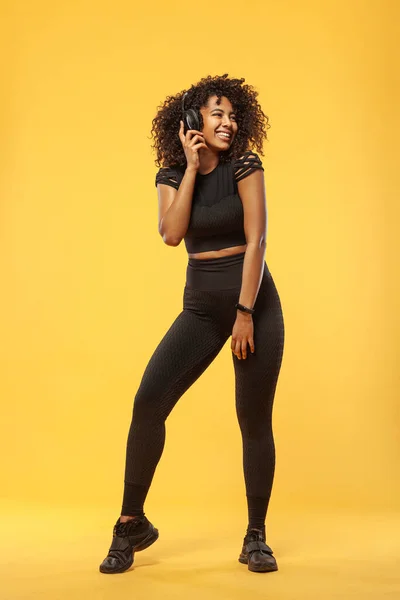 Concepto de estilo de vida. Retrato de una hermosa mujer afroamericana con el pelo rizado alegre escuchando música en el teléfono móvil. Fondo de estudio amarillo . — Foto de Stock