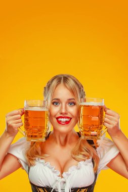 Geleneksel bir Bavyera ya da Alman dirndl 'i giymiş genç seksi Ekim Festivali kız garsonu, sarı arka planda içkili iki büyük bira bardağı servis ediyor..