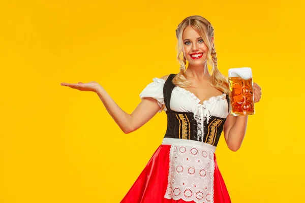 Garçonete jovem sexy Oktoberfest menina, vestindo um dirndl tradicional bávaro ou alemão, servindo caneca de cerveja grande com bebida isolada no fundo amarelo. Mulher apontando para a esquerda . — Fotografia de Stock