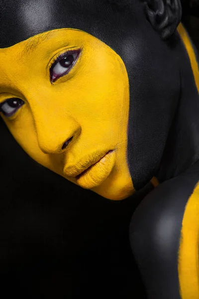 Arte facial. Mulher com tinta corporal preta e amarela. Menina africana nova com pintura corporal colorida. Um modelo incrível com maquiagem amarela. Cara fechada. — Fotografia de Stock