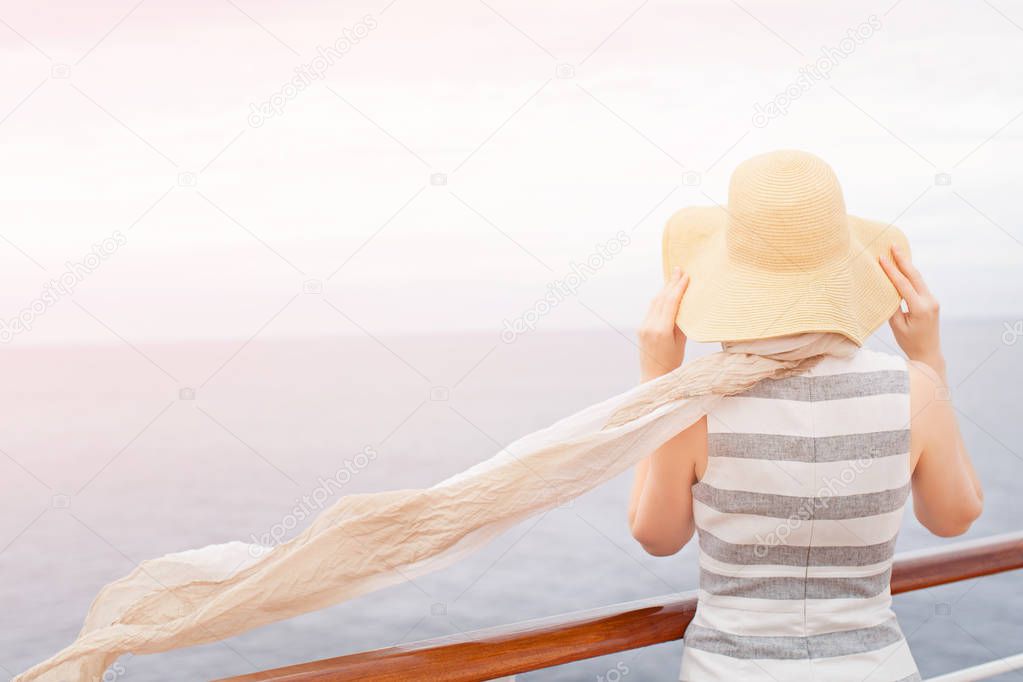 back view of young woman wearing sunhat and scarf waving on the wind enjoying cruise vacation looking at the sea, copy space on left, sun glare