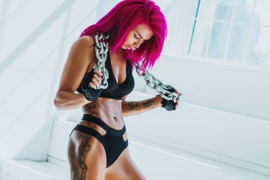 Parlak beyaz iç ağır zincir ile pembe saç ayakta olan genç fitness kadın. Vücudunda dövme.
