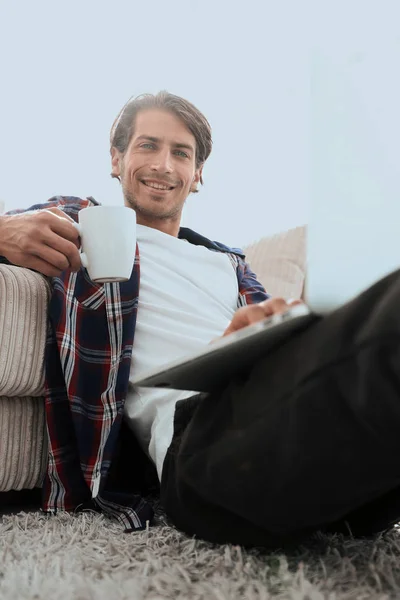 Genç adam kanepede yakın katta oturan bir fincan tutan dizüstü bilgisayar ile — Stok fotoğraf