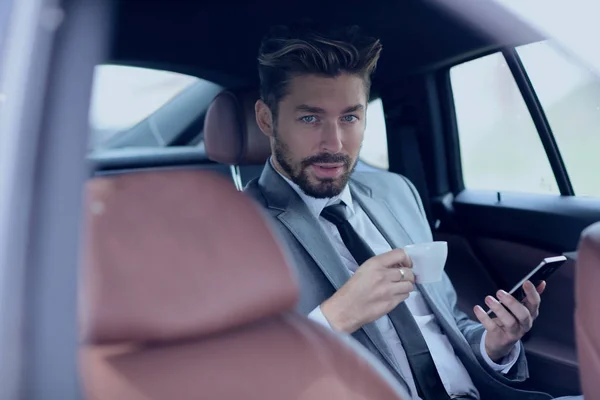 Uomo d'affari che beve caffè in auto con il telefono in mano — Foto Stock