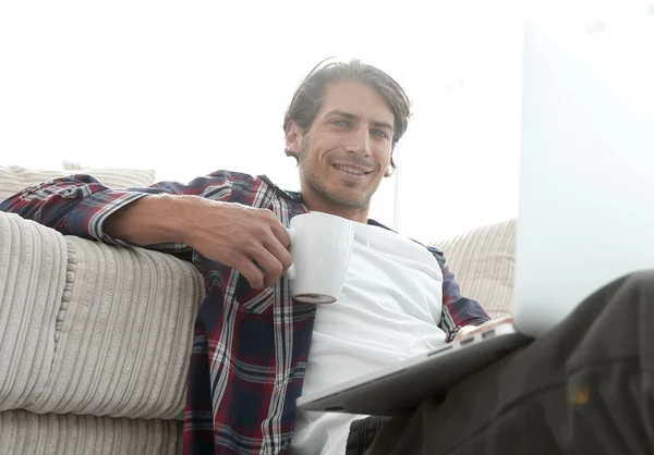 Young man with laptop holding a cup sitting on the floor near the sofa — Stock Photo, Image