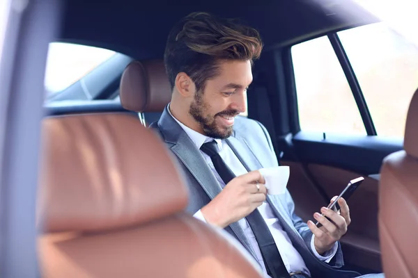 Podnikatel pije kávu v autě s telefonem v ruce — Stock fotografie