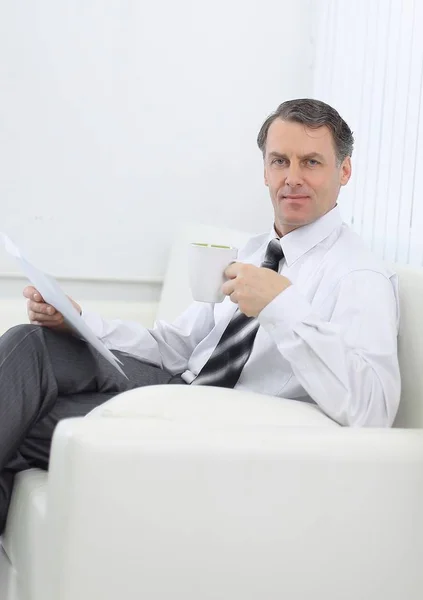 Hombre de negocios confiado bebiendo té y leyendo un documento, sentado en una silla en la habitación del hotel —  Fotos de Stock
